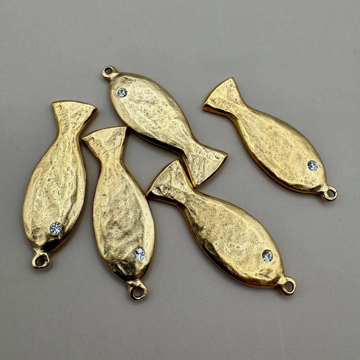 Close-up of five gold fish charms showcasing rhinestone eyes and attachment loops on a gray background