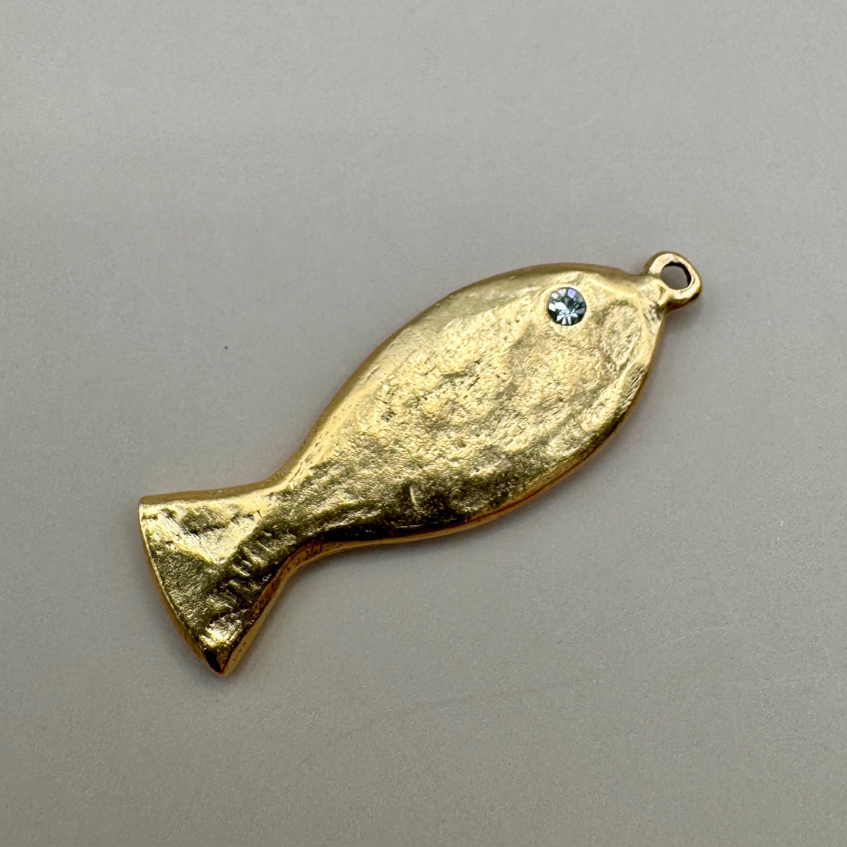 Angled view of a gold fish charm showcasing a rhinestone eye and top loop on a gray background